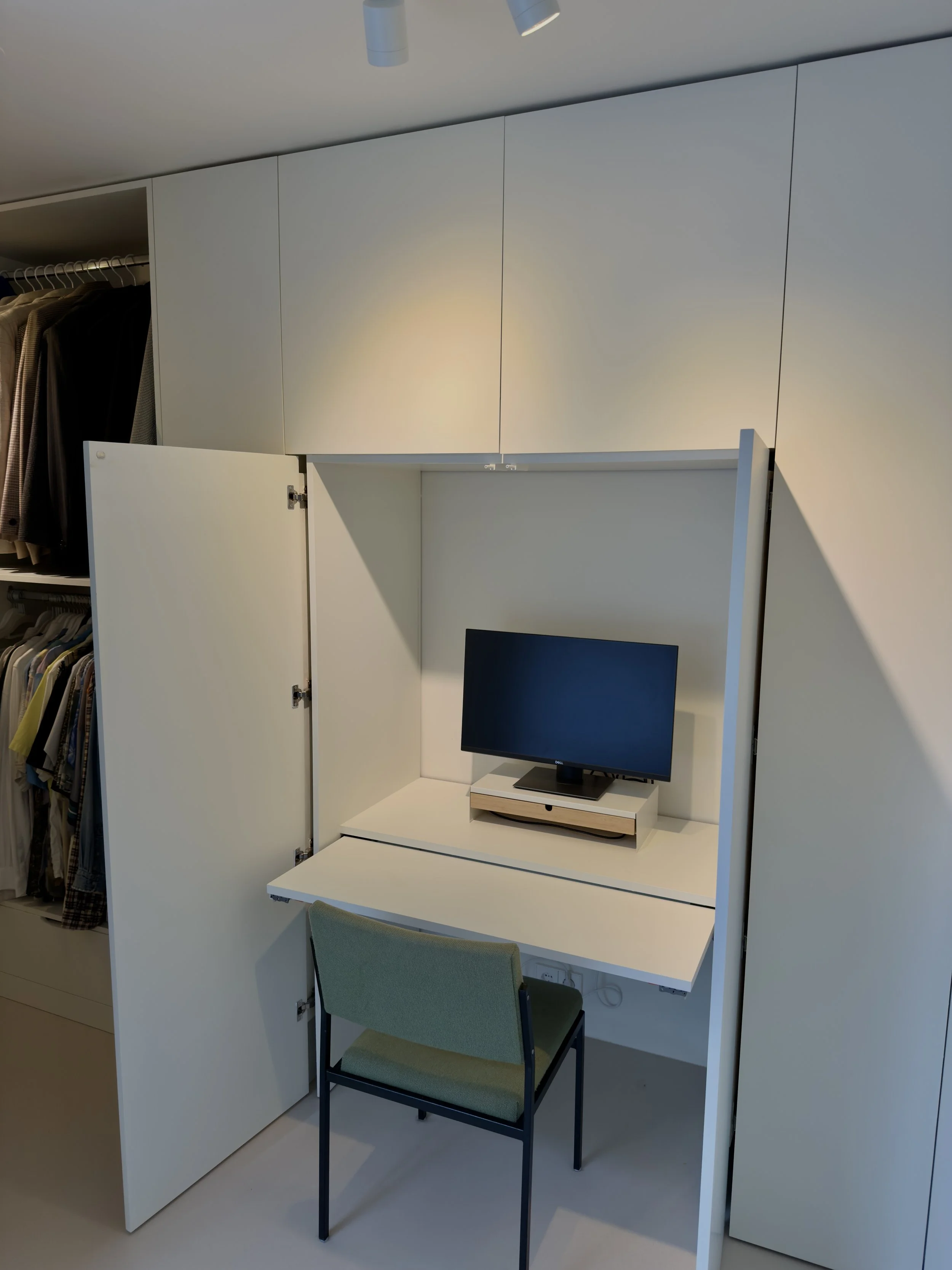 Een kleine werkplek met een opvouwbaar bureau, een groene stoel, en een computer met een monitor, ingebouwd in een witte kast in een kledingwinkel.