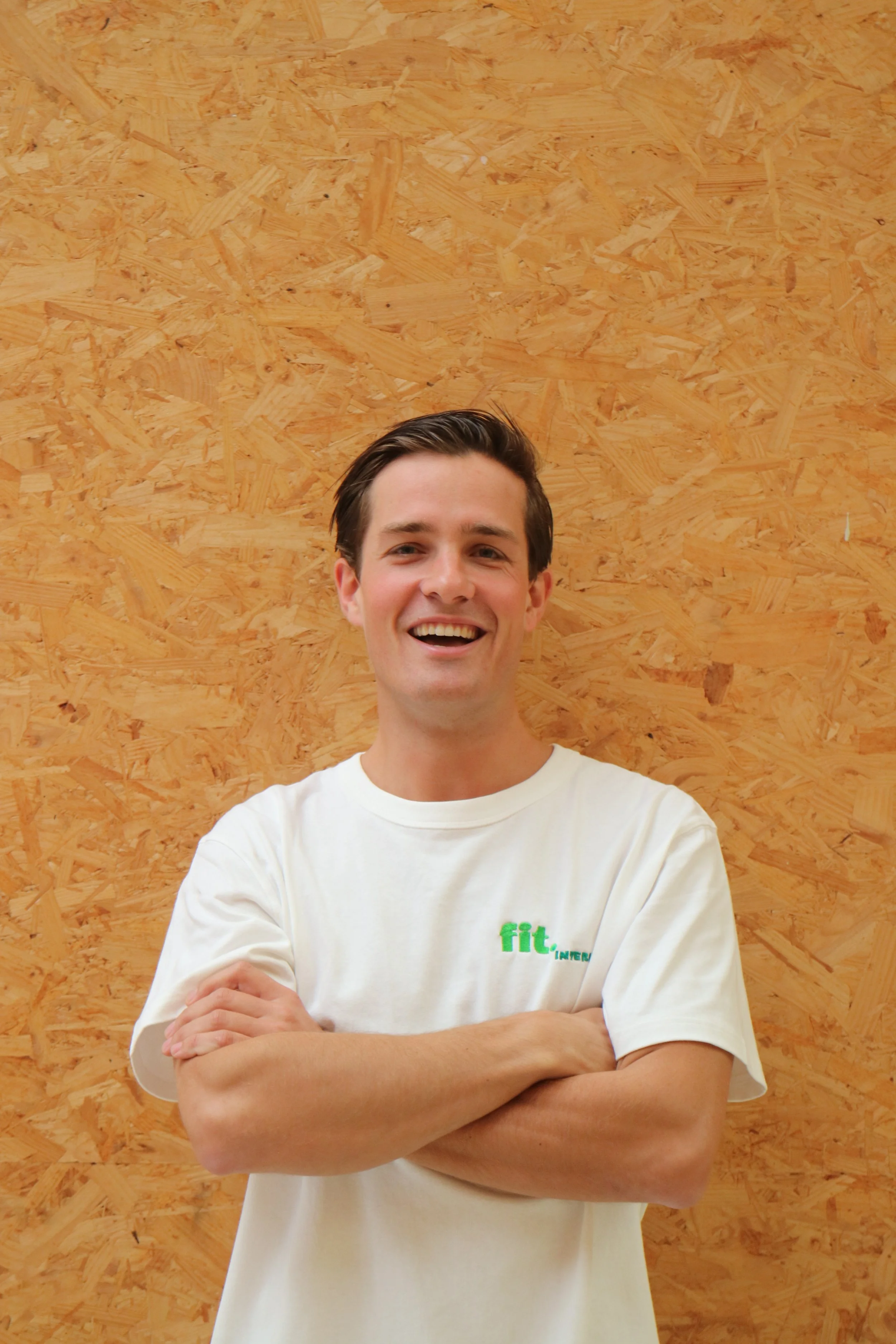 Een lachende man in een wit T-shirt met groen logo tekst 'fit. interior', staand voor een houten achtergrond.
