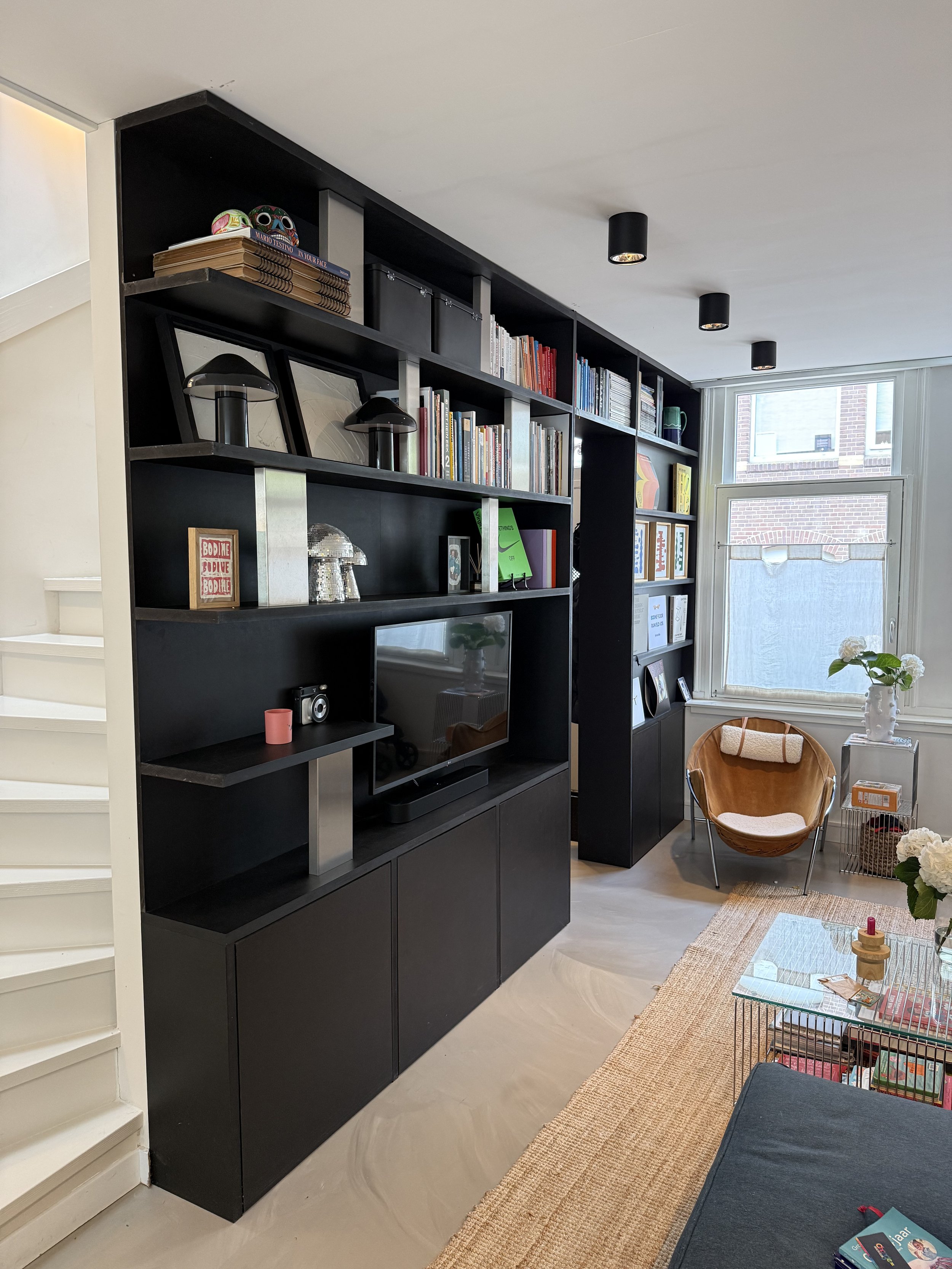 Interieur van een woonkamer met zwarte boekenplank, tv, planten, stoel, en raam.