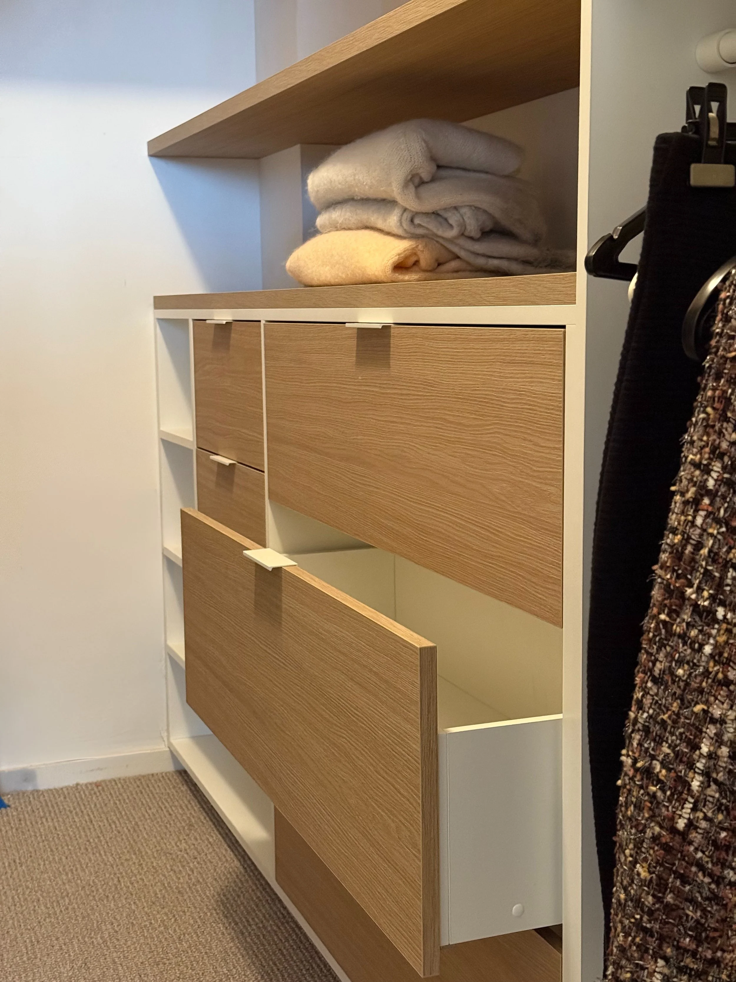 Een houten en witte kledingkast met open en gesloten lades, met gevouwen wollen dekens op de plank erboven, een kledingstuk hangt aan een haak aan de rechterkant.