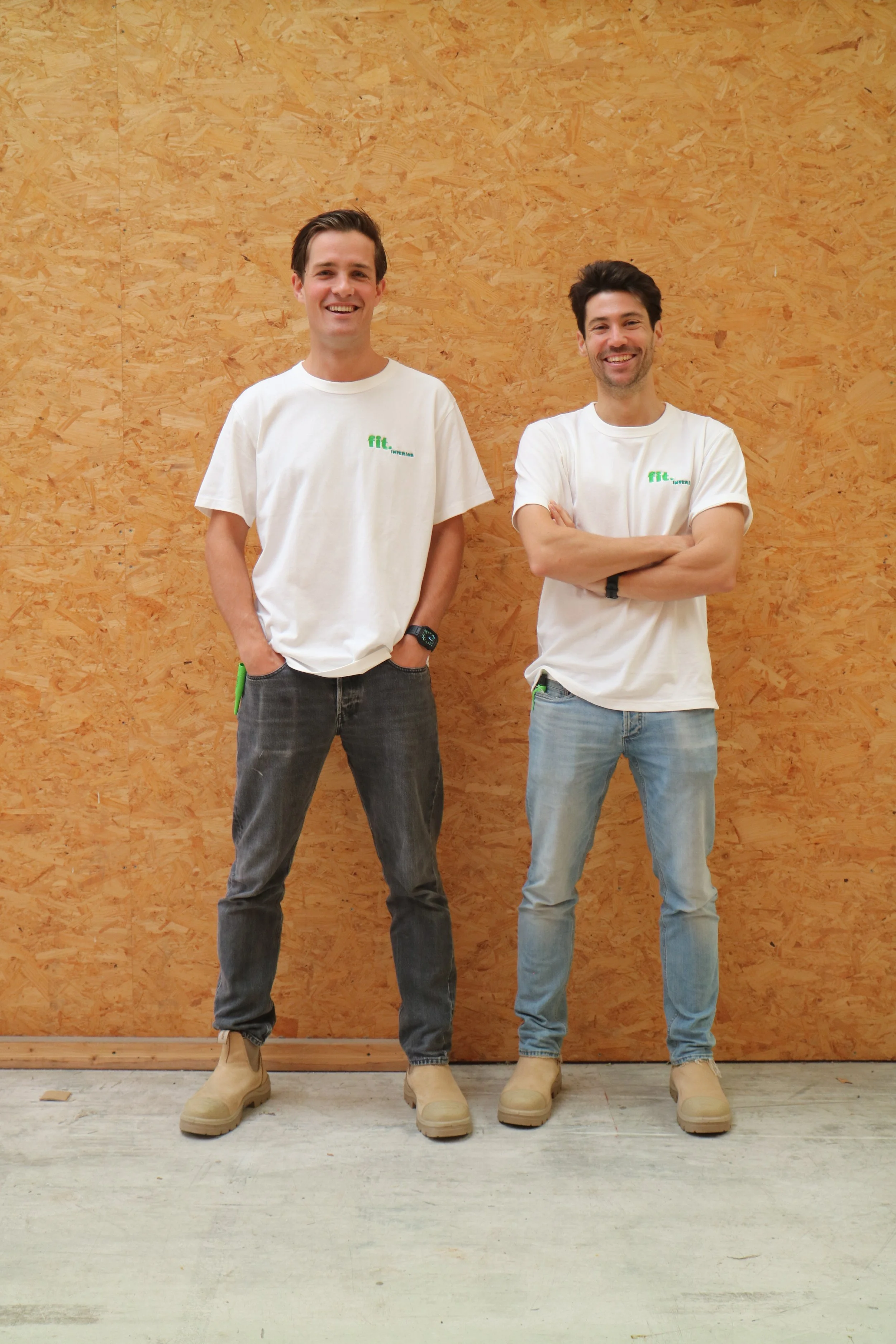 Twee jonge mannen staan naast elkaar voor een houten muur, beiden dragen witte T-shirts met groene tekst en dragen beige werklaarzen. Ze lachen en kijken naar de camera.