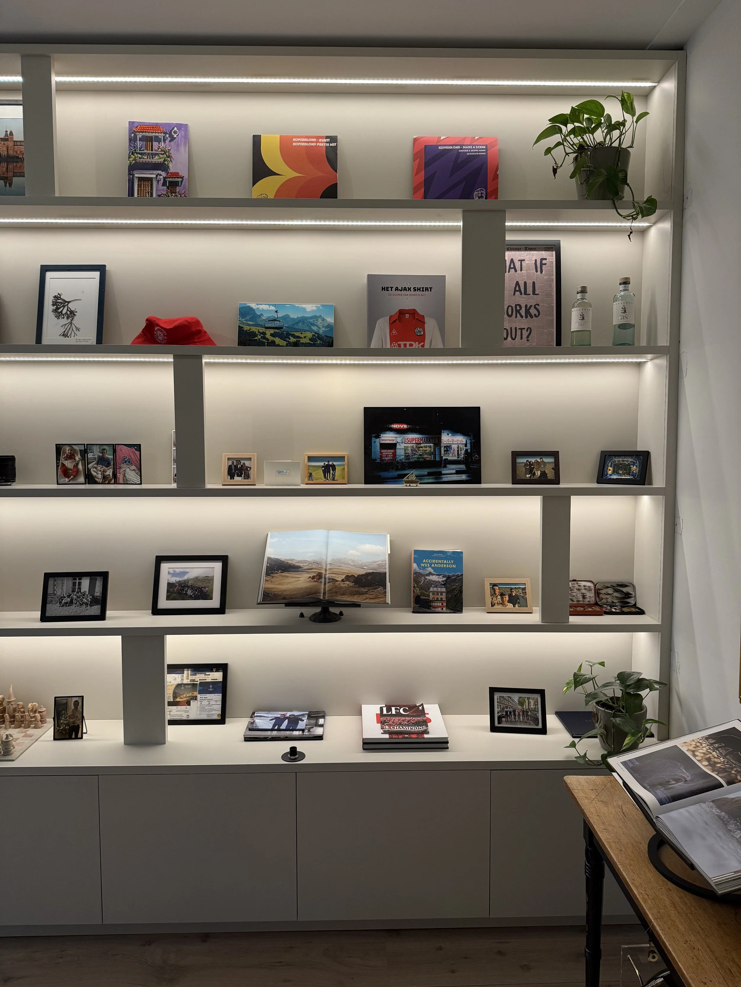 Witte boekenplank met foto's, boeken en decoraties, verlicht met ingebouwde LED-verlichting, met een tafel op de rechterkant met een open boek en een plant.