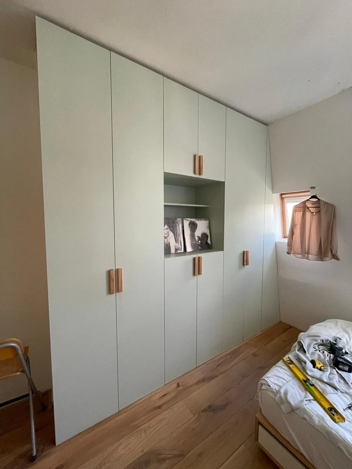 Grote witte kast met houten handvatten in een kamer met houten vloer, een klein raam met een jas er aan gehangen, en een bed met gereedschap erop.