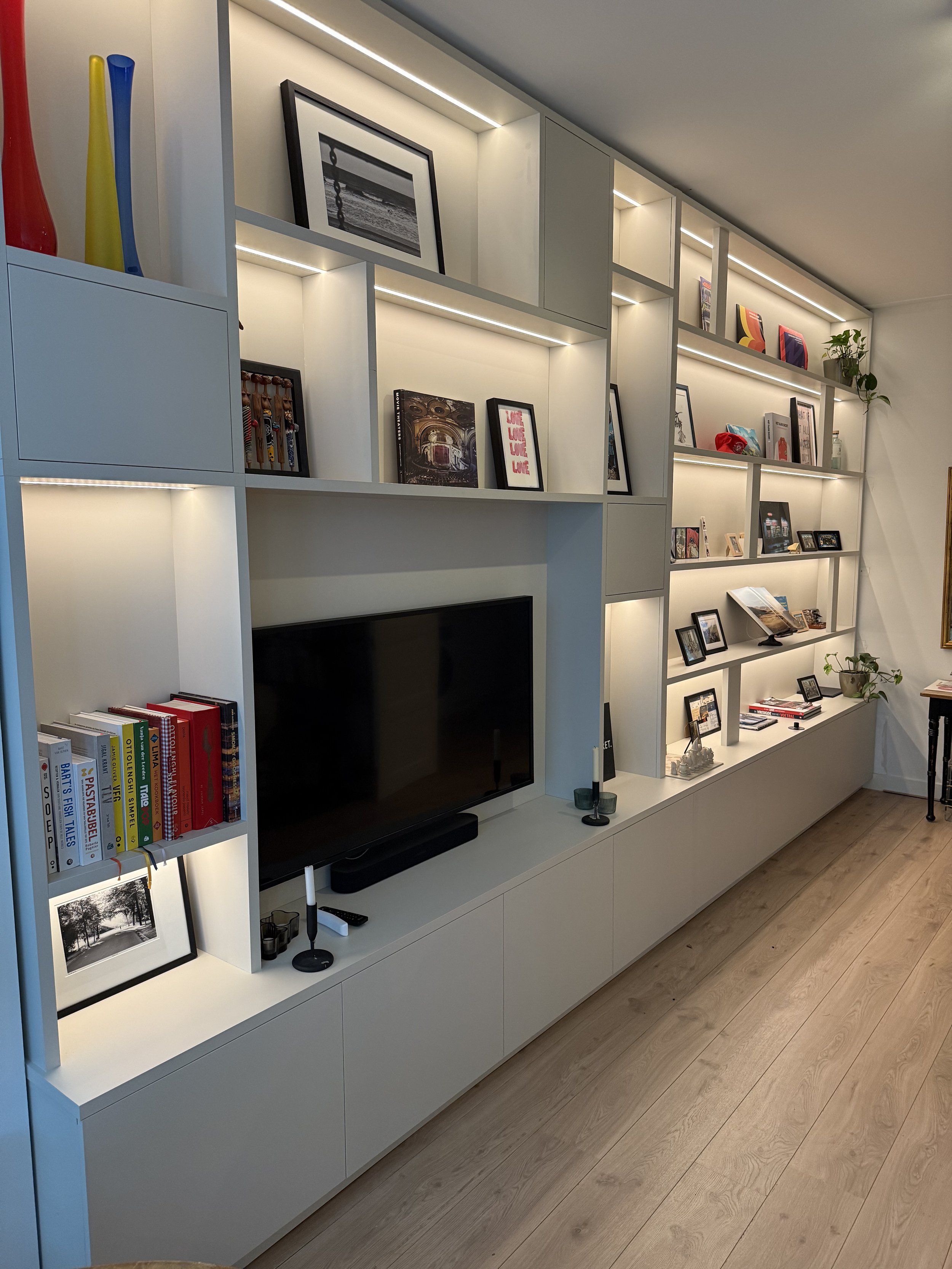 Witte wandrekken met boekstapels, foto’s, decoraties en een televisie, op een houten vloer.