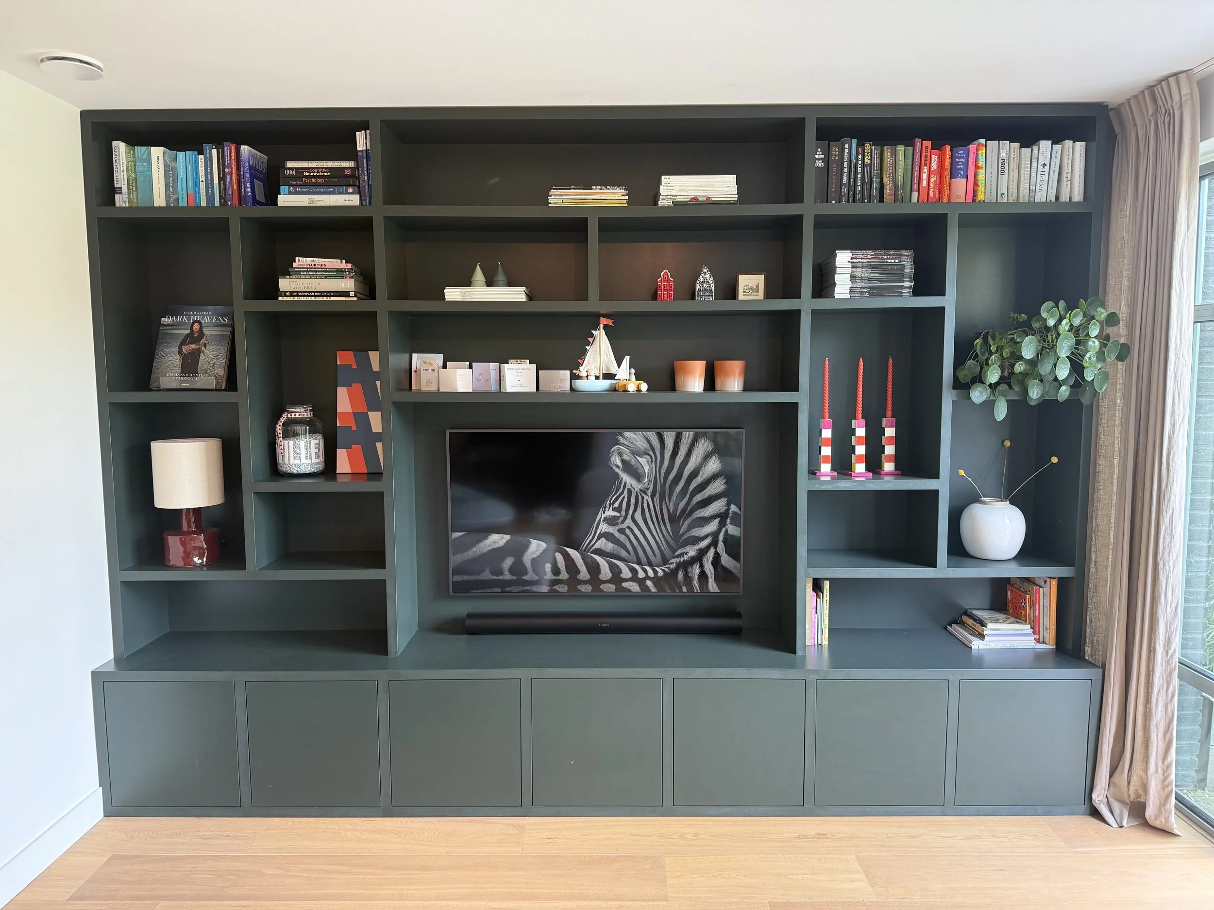 Groene boekenwand met boeken, decoraties, een tv met een zebra-afbeelding, en planten, naast een raam met gordijnen.