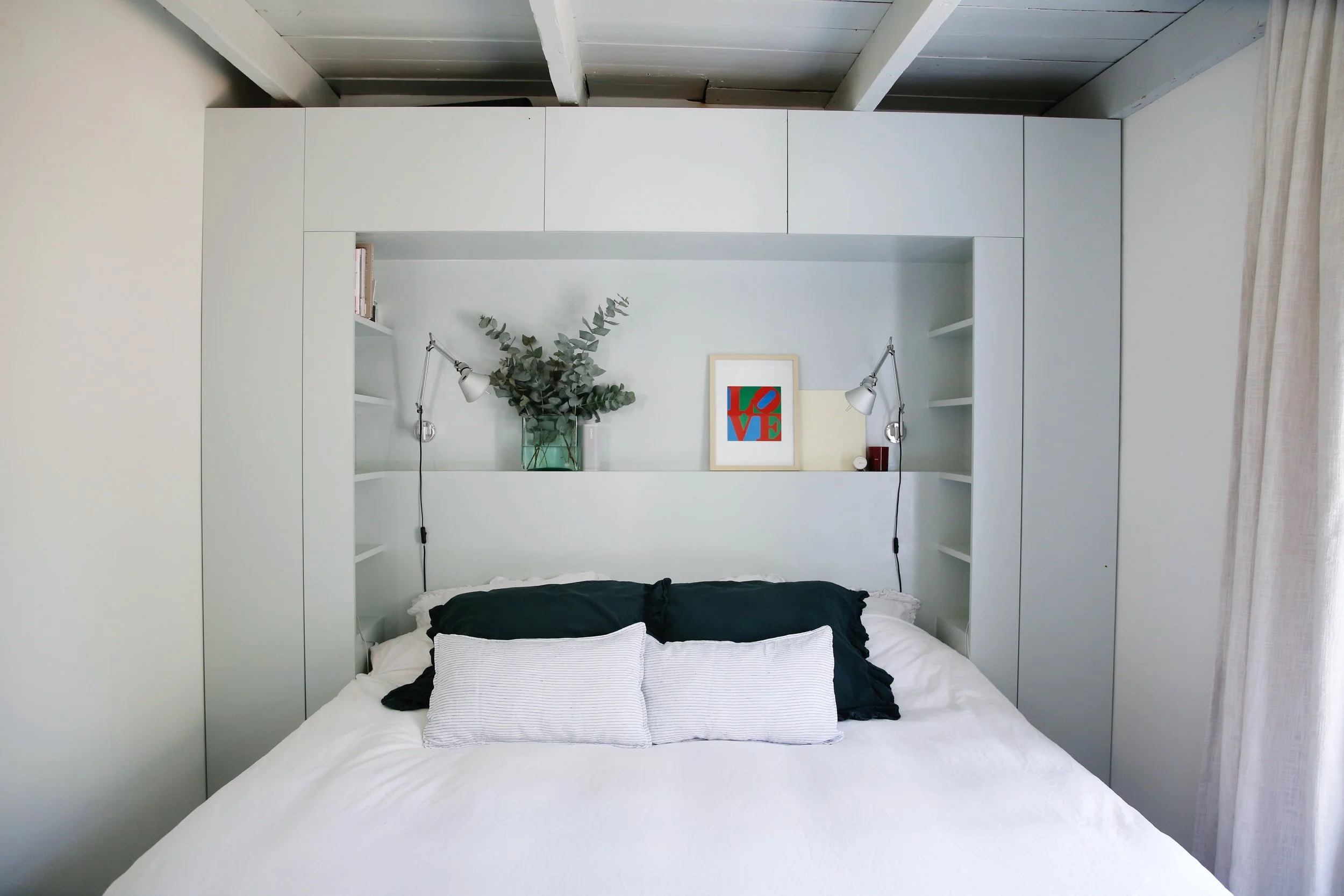 Slaapkamer met wit bed, zwart en witte kussens, ingebouwde witte boekenkasten achter het bed, decoratie met een vaas met groene bladeren, een geframed kunstwerk met de tekst 'LOVE', twee wandlampen, een raampartij met witte gordijnen.