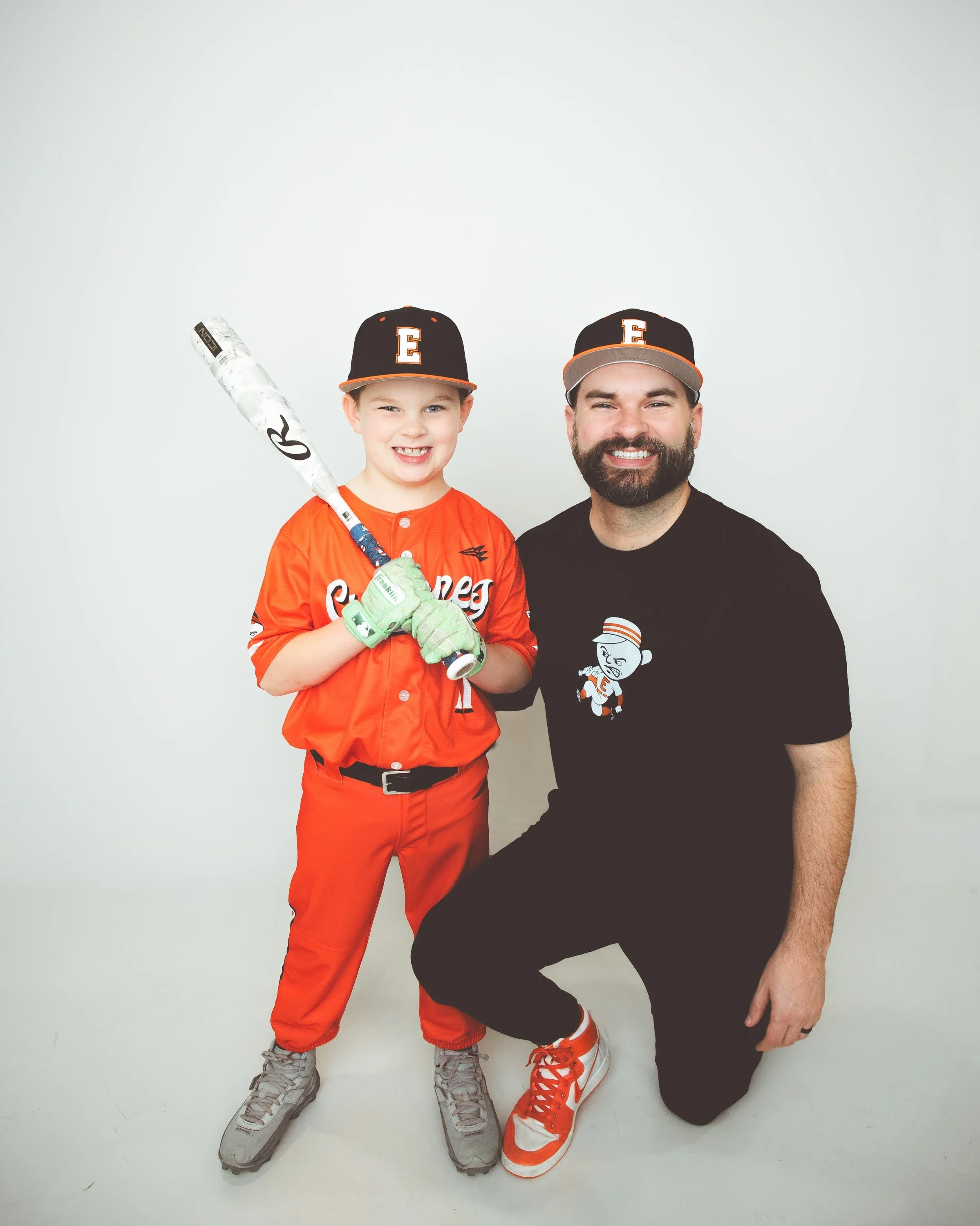 A man and a young boy in baseball uniforms on a baseball field, holding a baseball and a glove, smiling at the camera.