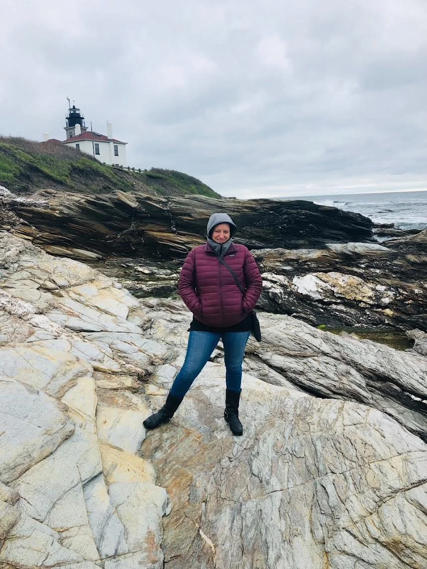 A woman standing on rocky terrain near the coast, wearing a purple jacket, jeans, and a gray hoodie, with a lighthouse and cloudy sky in the background.