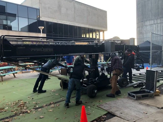 A film crew working on a set outdoors, with a camera rig and equipment, in a city area with modern buildings and a green ground surface.