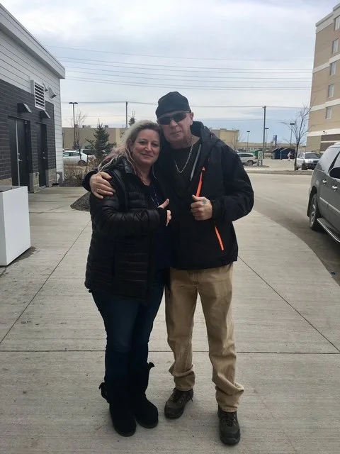 Two people, a woman and a man, standing together on a sidewalk outside, with their arms around each other, smiling at the camera.