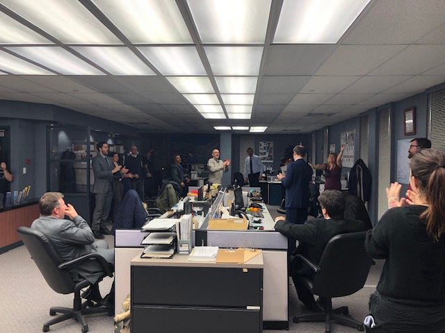 Business meeting in a corporate office with people standing and sitting, some clapping, during a presentation or announcement.