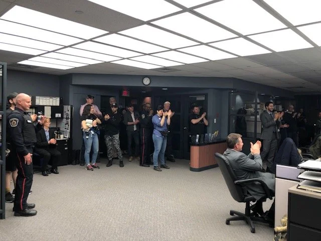Group of people, including a police officer, standing and clapping in an office setting, with some seated at desks and others gathered near the doorway.
