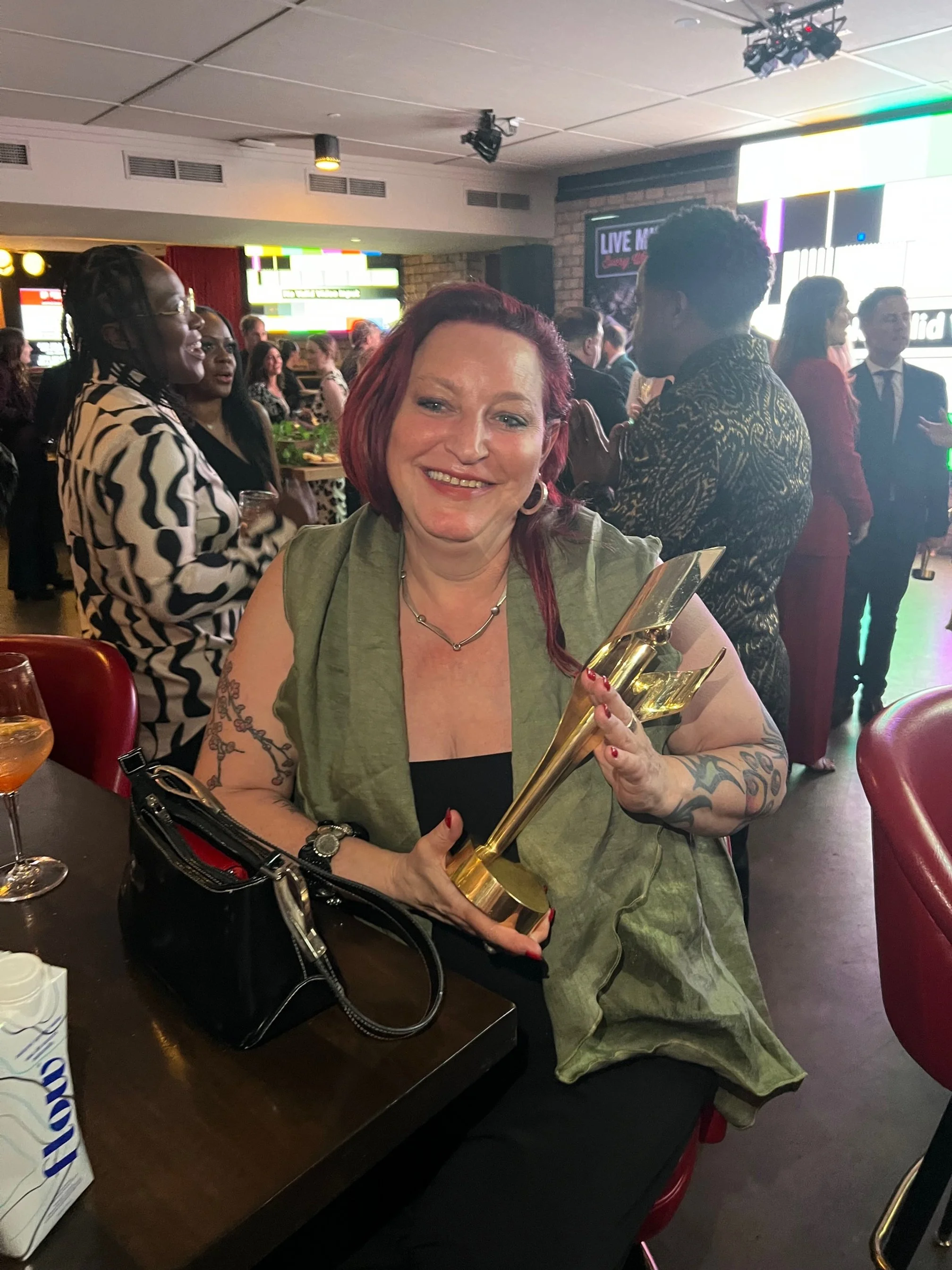 Woman with red hair and tattoos holding a golden award trophy, smiling at a celebration event with people in the background.