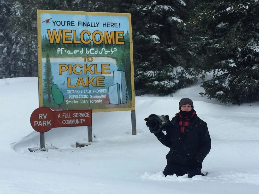 A person standing in snow next to a colorful welcome sign that reads "You're finally here! Welcome to Pickle Lake. Ontario's last frontier. Population: somewhat smaller than Toronto." The sign also indicates it's an RV park and full service community