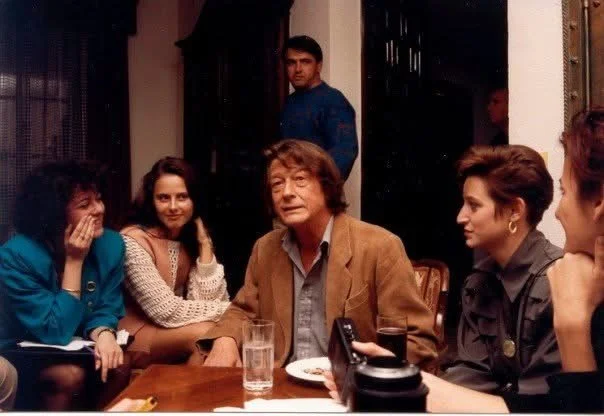 Group of six people seated around a table in a restaurant or cafe, with one person standing in the background.