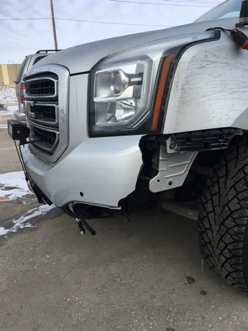 Close-up of the front left side of a silver pickup truck with visible damage to the bumper and wheel well.