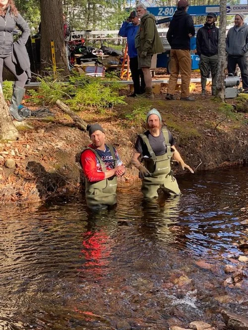 Two men wearing waders are standing in a river, surrounded by a wooded area, with a group of people and equipment on the riverbank in the background.