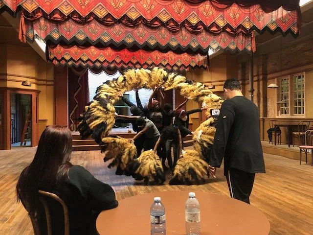 Group of performers practicing a drum routine on a stage with a large feathered hoop while an audience member records with a smartphone.