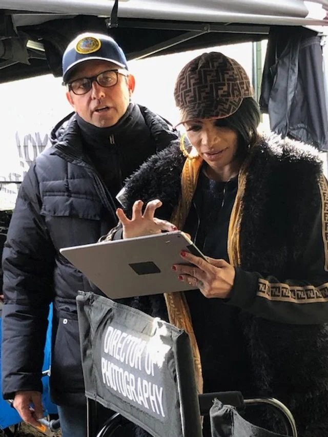 A man and woman are standing together outdoors, looking at a tablet. The woman is wearing a patterned cap, large hoop earrings, and a patterned jacket, while the man is wearing glasses and a cap with a yellow emblem. There is a sign that reads "DIREC