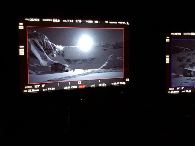 Nighttime shot of a mountain landscape with snowy peaks and a cabin, visible on the camera monitor.