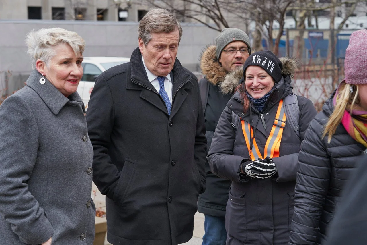 A group of five people standing outside in cold weather, wearing winter coats and accessories, engaged in conversation.