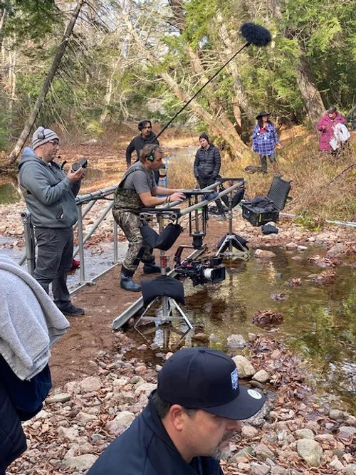Film crew filming a scene by a creek in a wooded area with equipment, crew members, and actors.