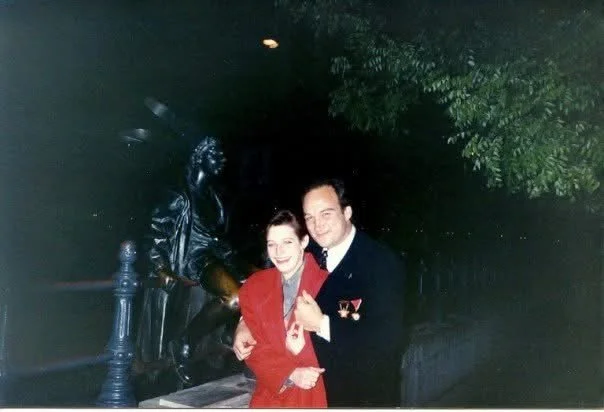 A man in a dark military uniform with medals and a woman in a red dress, standing close together and smiling outdoors at night.