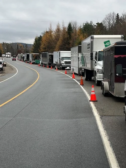 Line of trucks and vehicles parked on the side of a curved road, with orange traffic cones separating them, in a rural area with trees in the background.