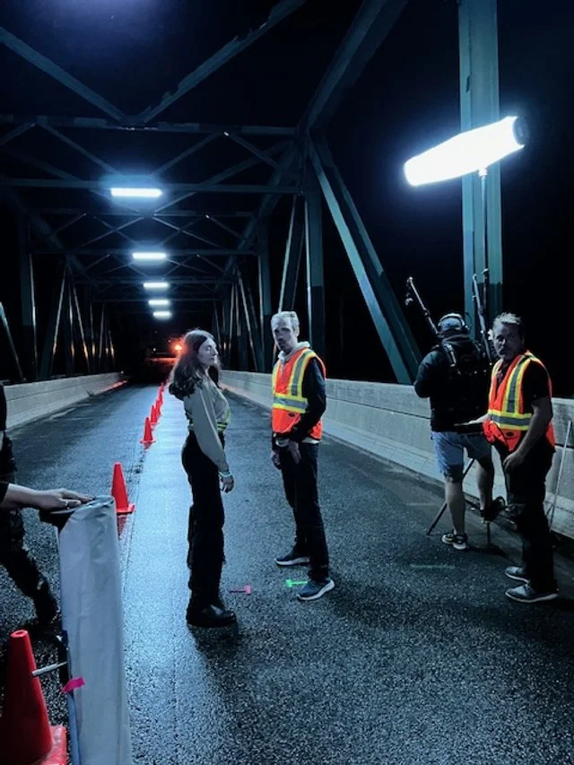 People standing on a dark bridge at night, with some wearing safety vests, next to production equipment including lights and a reflector, possibly filming or working on a project.