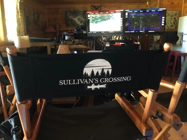 Film director's chair with the logo 'SULLIVAN'S CROSSING' and a forest design, facing two computer monitors on a wooden desk in a cozy, wood-paneled room.