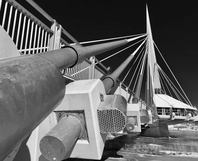 Black and white photo of a large fountain with multiple water jets shooting upward, situated on a concrete platform at a marina with sailboats in the background.