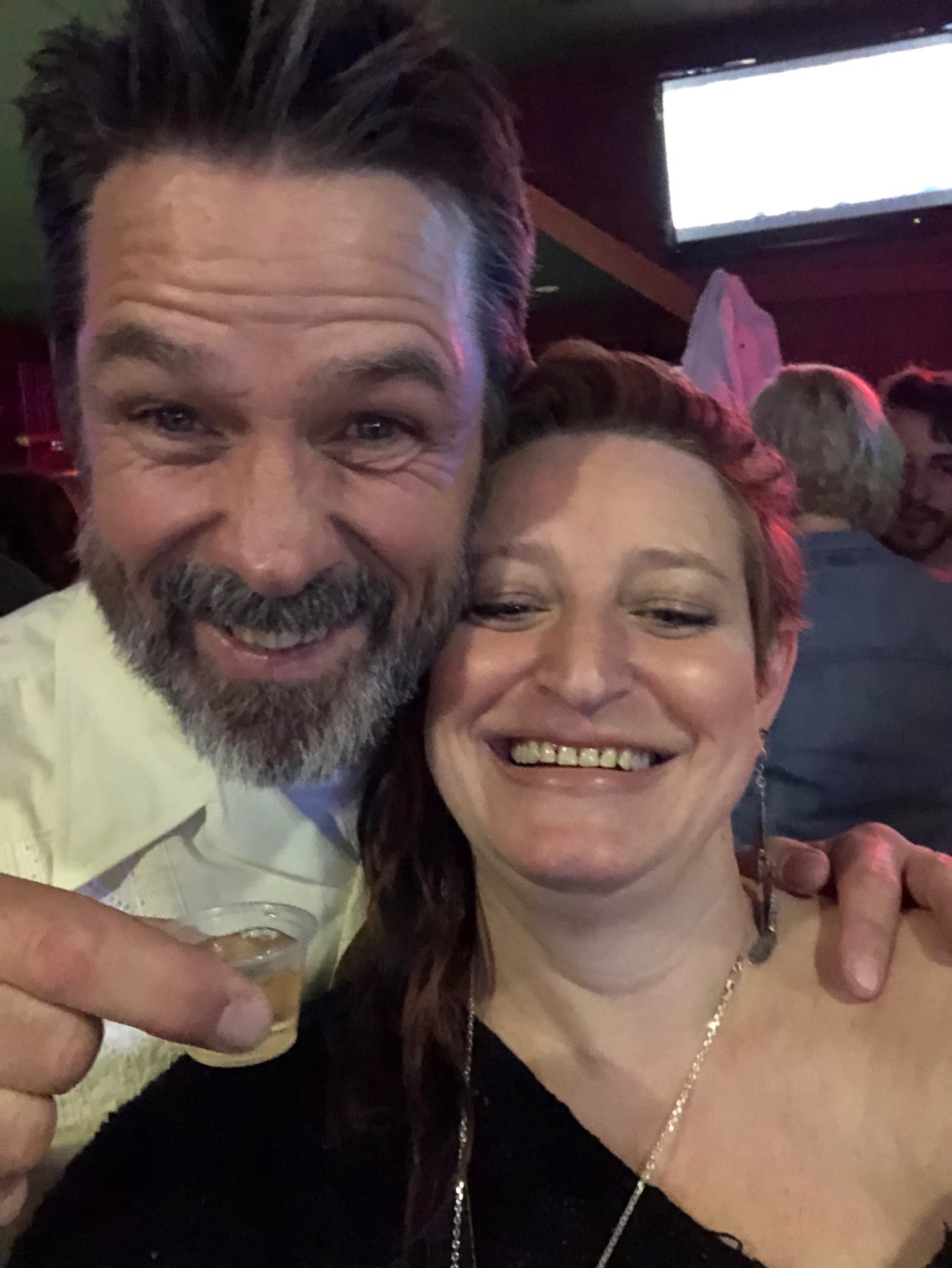 A man and woman smiling at a bar or social gathering, with the man holding a shot glass. The background shows other people and a television screen.