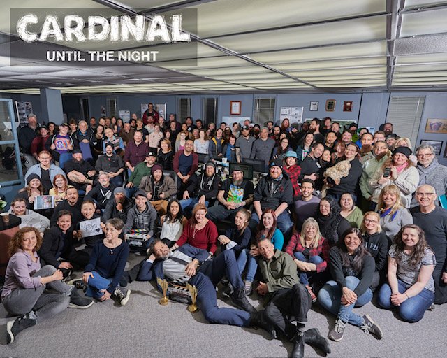 Large group of people gathered in an indoor space for a group photo, with a sign that reads 'Cardinal Until the Night' hanging from the ceiling.