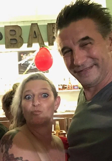 A man and a woman making funny expressions for a selfie in a bar or restaurant, with the word 'BAR' visible in the background and a red balloon hanging from the ceiling.