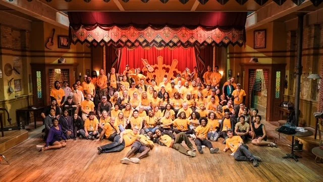Large group of people, some wearing yellow shirts, gathered on a wooden stage with a red curtain and musical instruments in the background, posing for a group photo.