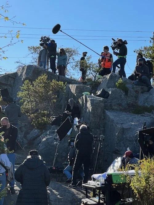 A film crew filming across a rocky outdoor landscape, with equipment, crew members, and camera operators, some in jackets and masks, under a clear blue sky.