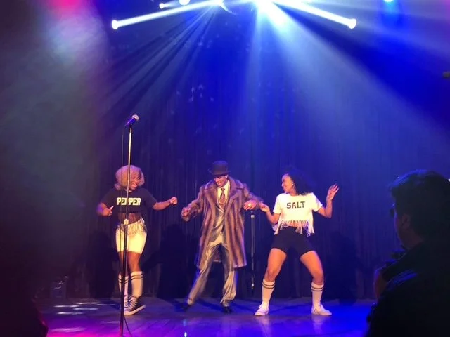 Three performers dancing on stage under colorful lights with a dark curtain background, one man in a striped suit and bowler hat, and two women in casual outfits.
