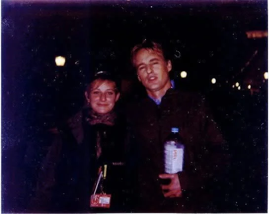 Two young men standing together in a dimly lit outdoor setting at night, one holding a water bottle, both smiling at the camera.