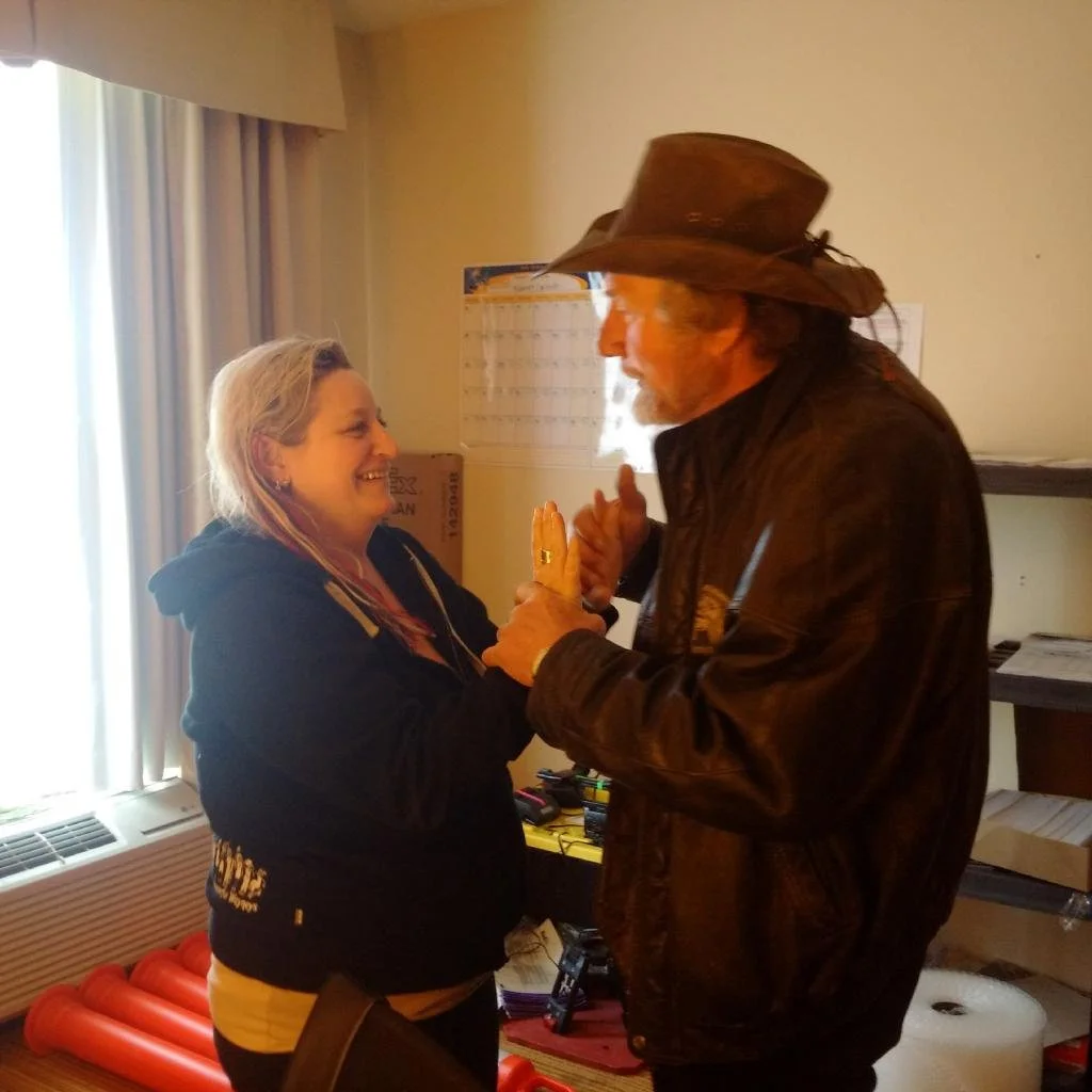 A woman and a man are smiling and holding hands inside a room. The woman has blonde hair and is wearing a black hoodie, while the man is wearing a brown leather jacket and a wide-brimmed hat. They appear to be engaged in a friendly or intimate conver