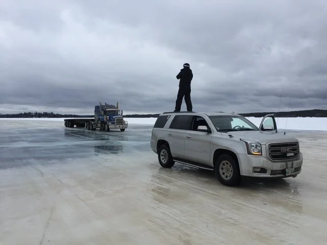 A man standing on top of a white SUV parked on a frozen lake, with a semi-truck and trailer in the background on the ice, under a cloudy sky.