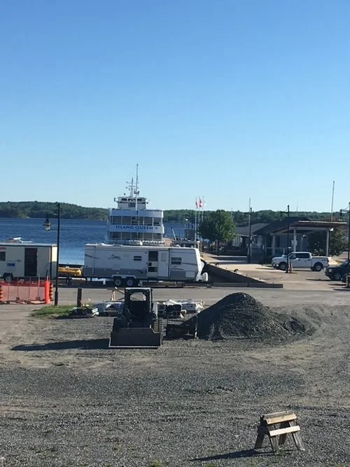 View of a waterfront with a large boat, labeled 'Island Queen,' docked near a small building and several parked cars. Construction equipment and gravel are in the foreground.