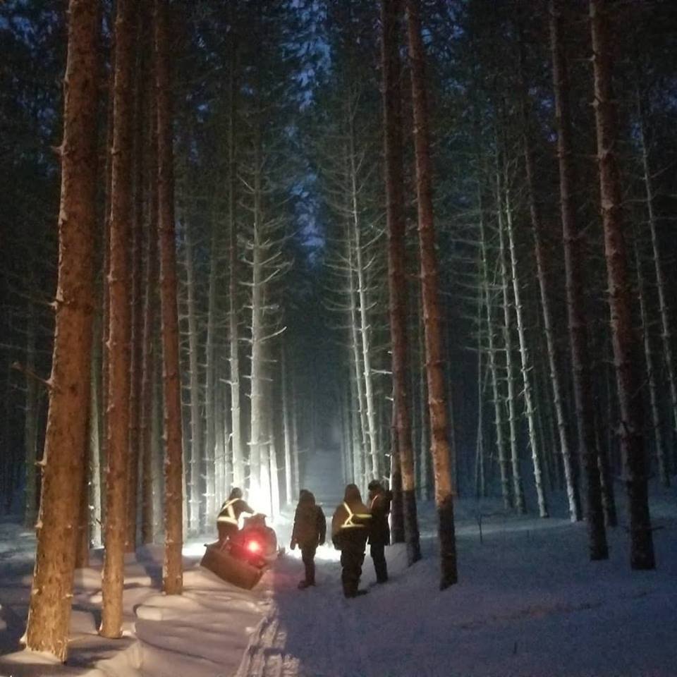 A group of four people walking through a snowy forest at night, illuminated by a bright light behind them, with a snowmobile on the ground nearby.