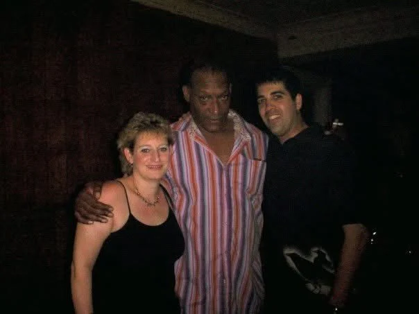 Three people posing together indoors, two men and one woman, with dark background.