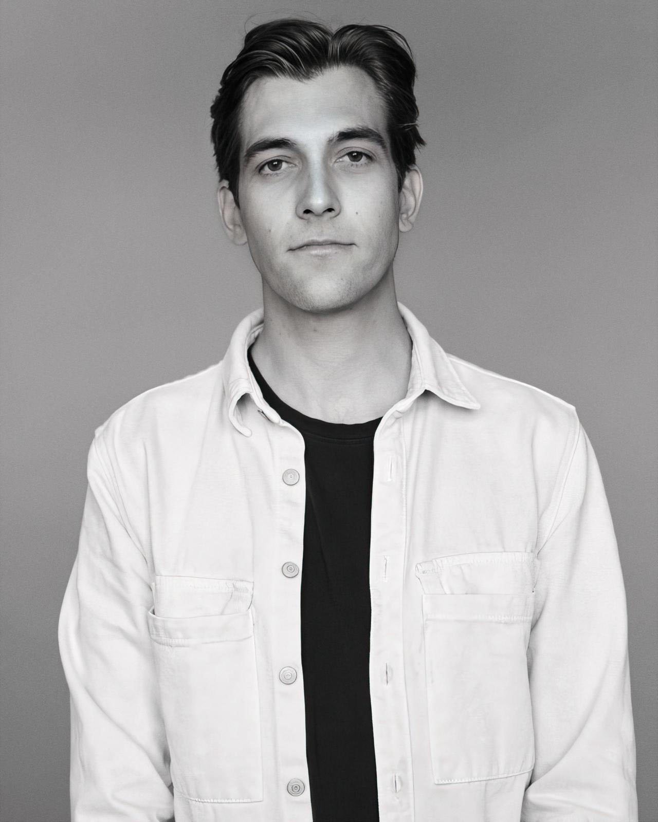 Black and white photo of a young man with dark hair wearing a light-colored button-up shirt over a dark T-shirt, standing against a plain background.