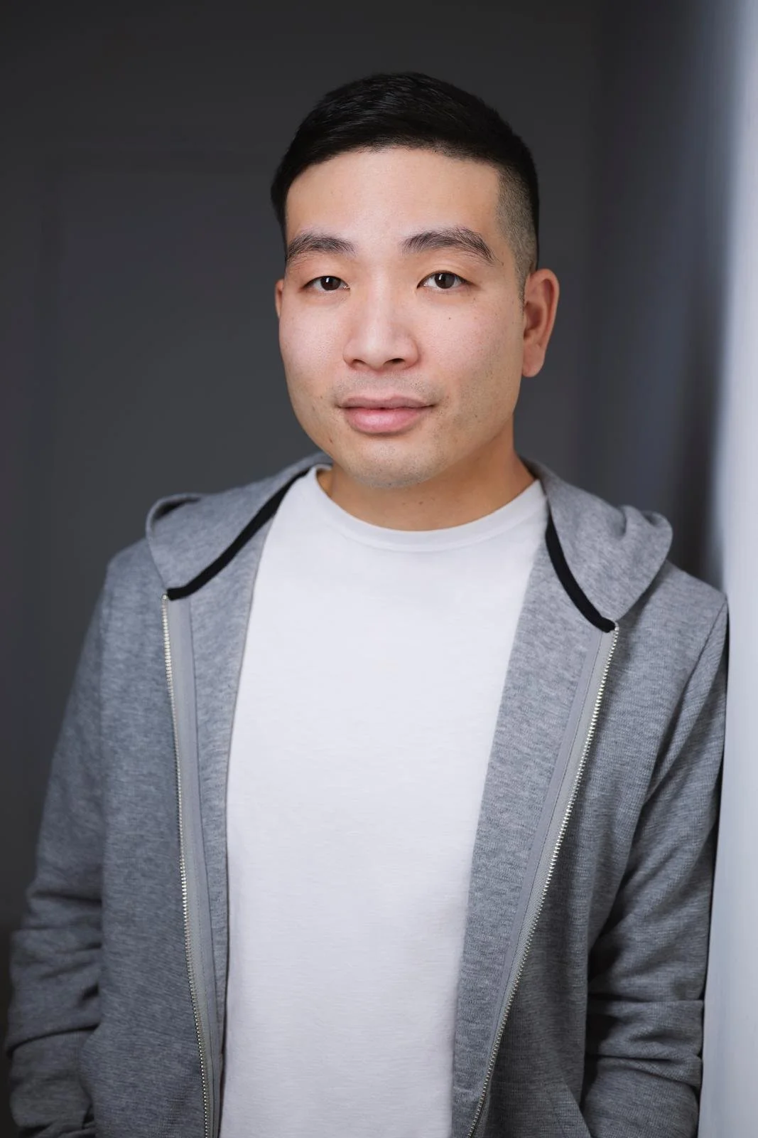 Portrait of a young man with short black hair, wearing a light gray hoodie over a white T-shirt, standing against a plain dark background.