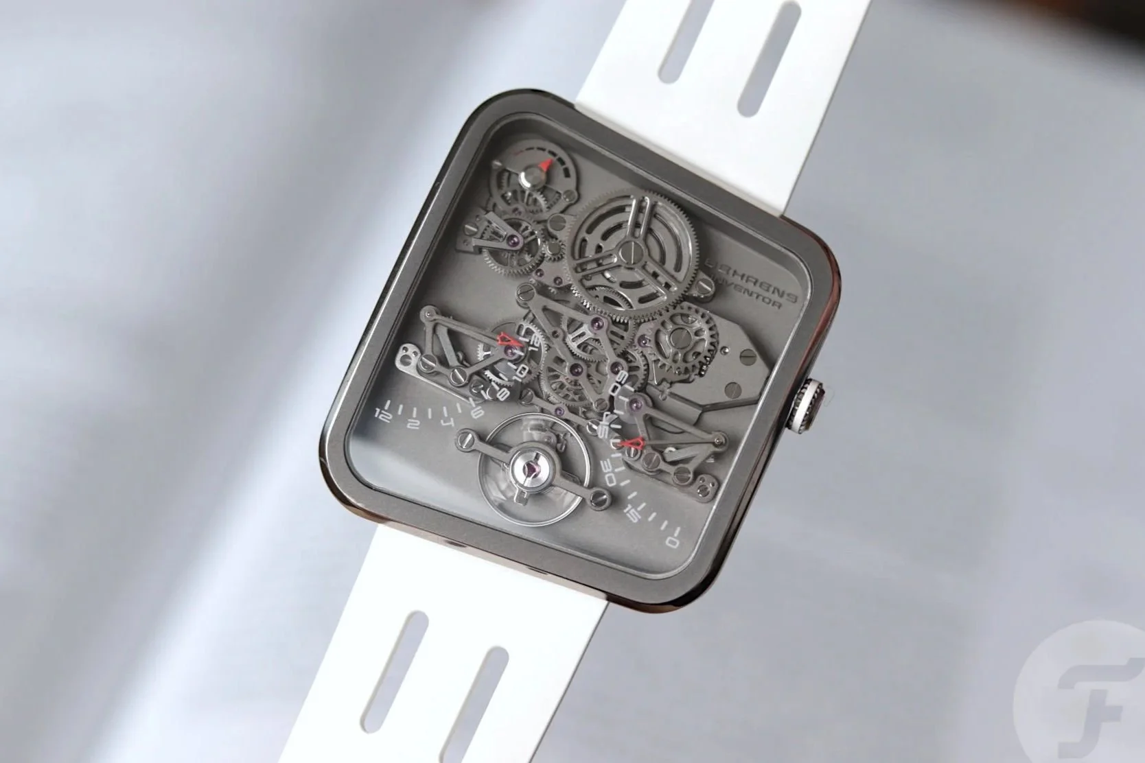 Close-up of a modern square watch with an open skeleton dial showing gears and mechanical parts, placed on a white strap against a plain background.