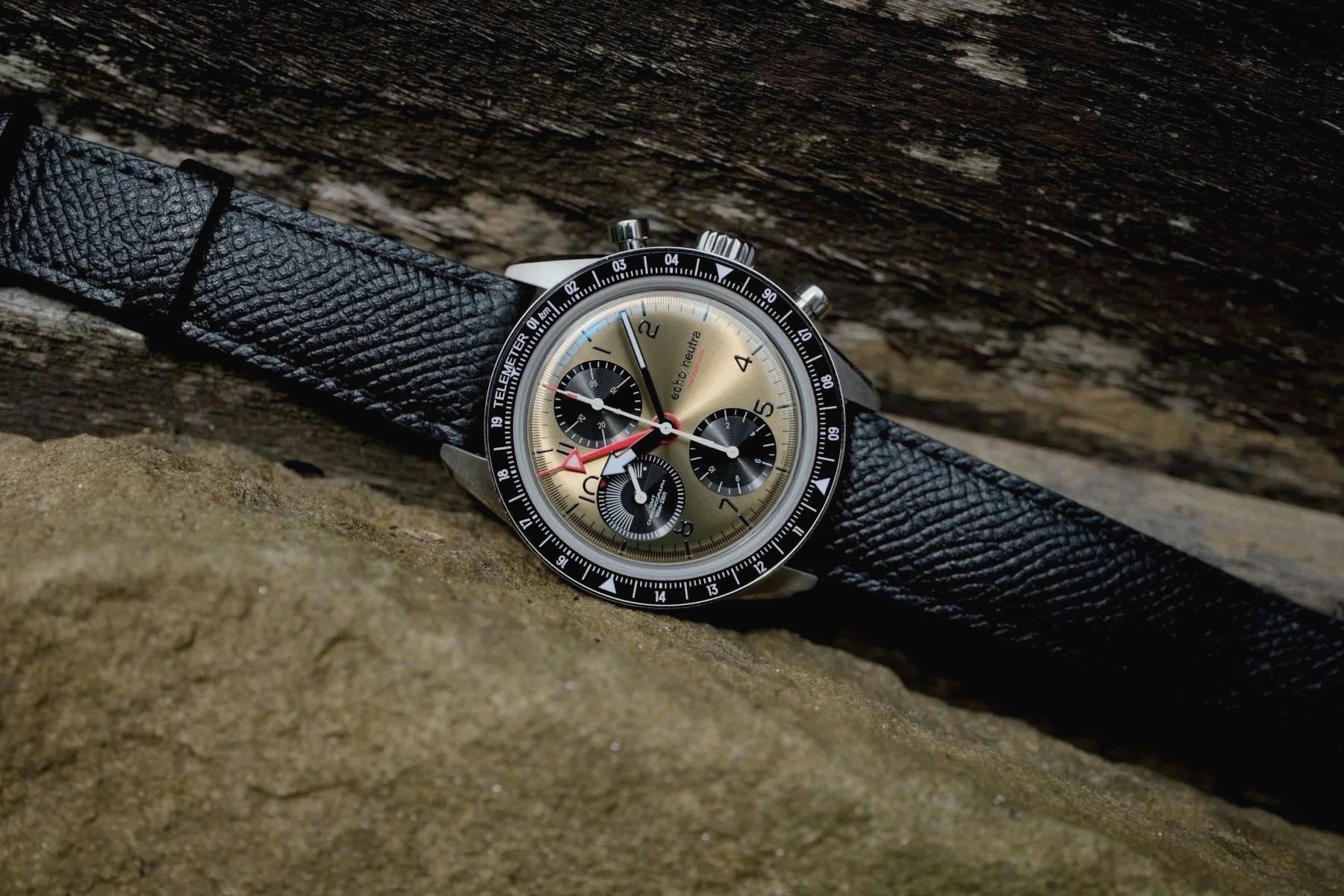 A wristwatch with a black leather strap and a gold-colored dial, resting on a rock surface with wooden background.