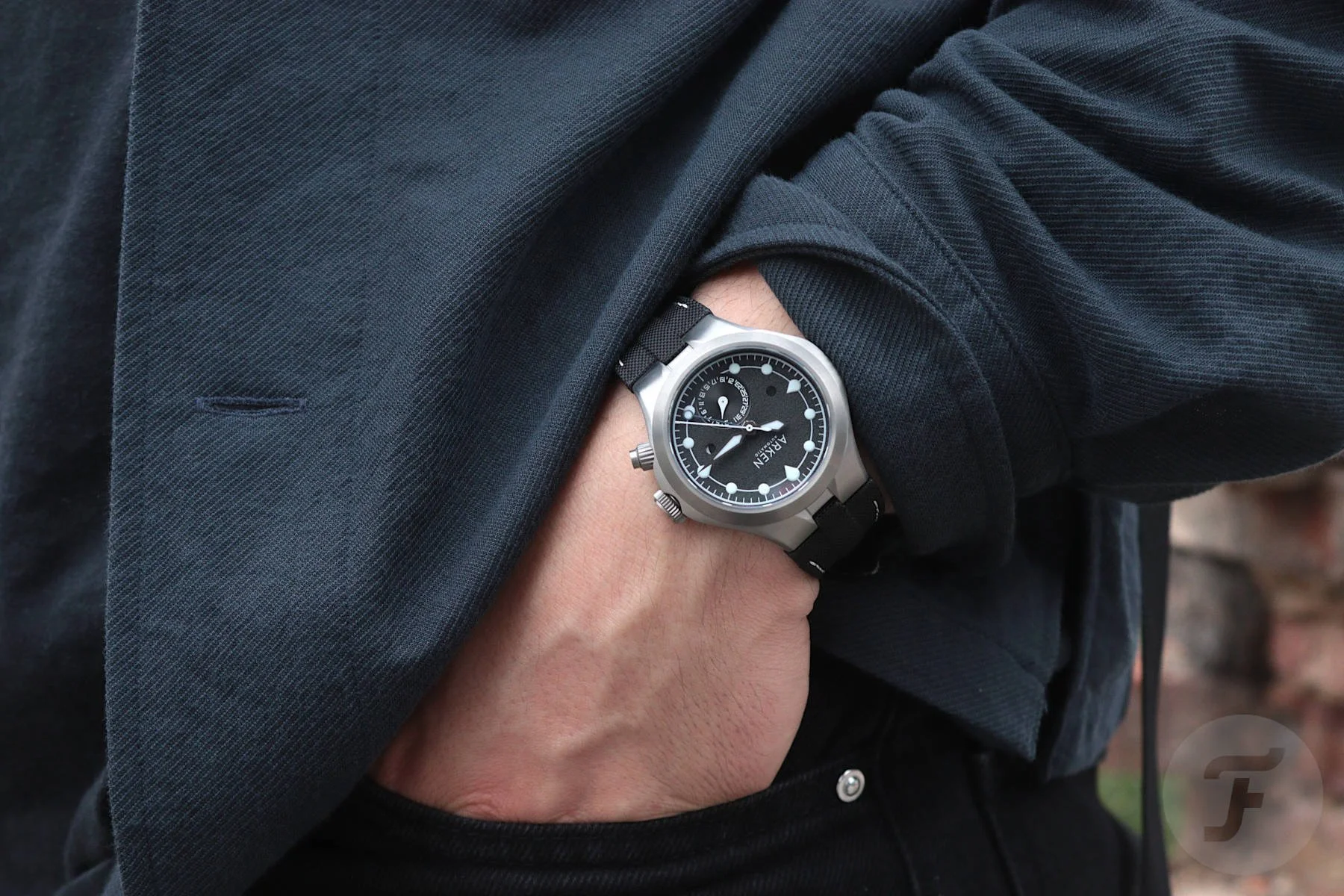 Person wearing a black and gray wristwatch with a black strap, with their hand in their pocket, dressed in a dark jacket and black shirt.