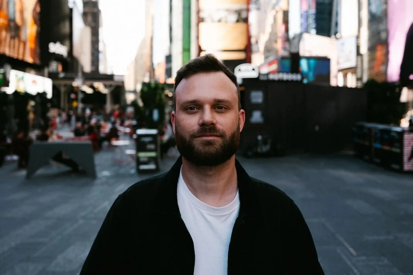 A man with a beard and short hair standing in Times Square in New York City, with tall buildings and billboards in the background.