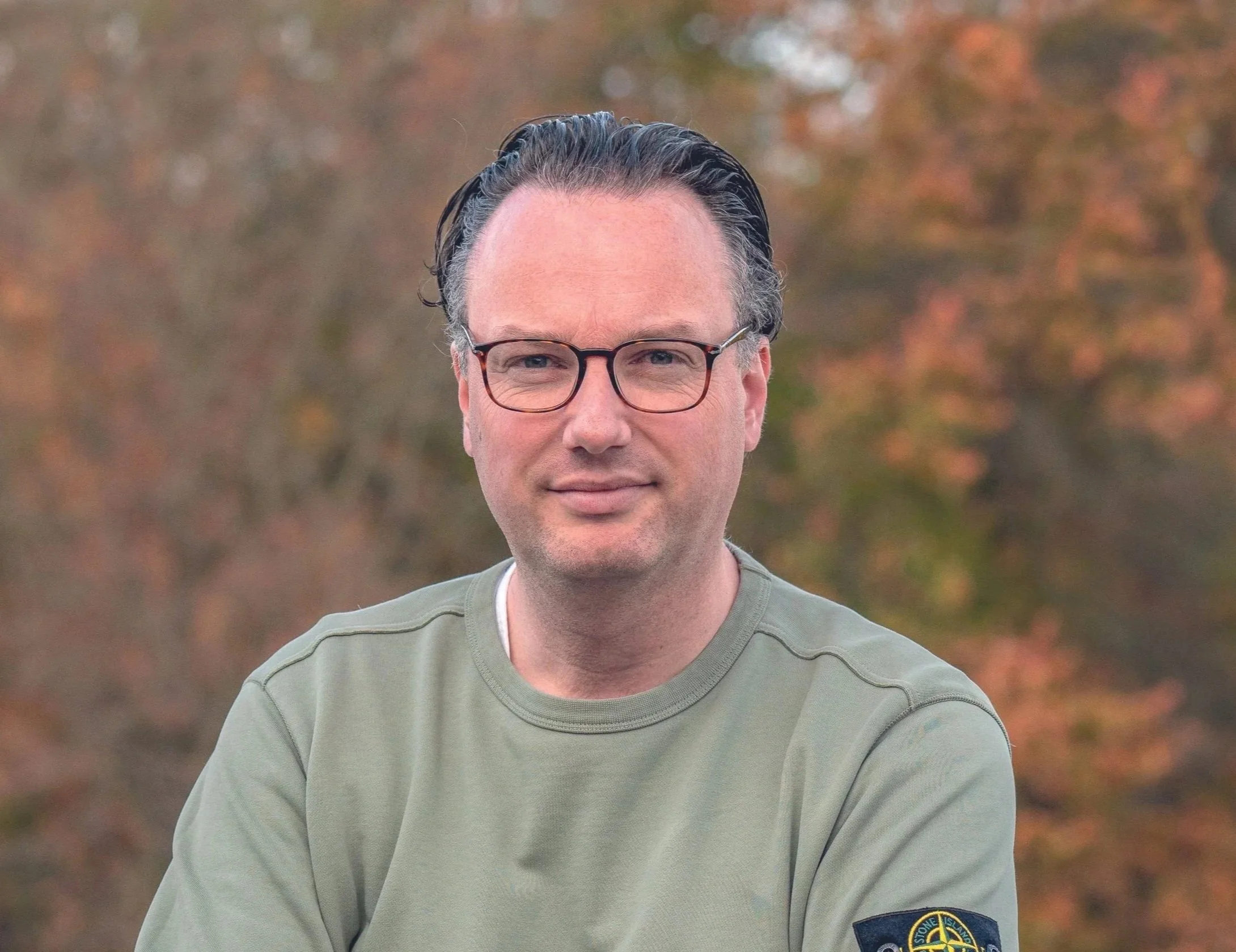 A man with glasses and dark hair is smiling outdoors with fall foliage in the background, wearing a khaki-green shirt with a patch on the sleeve.